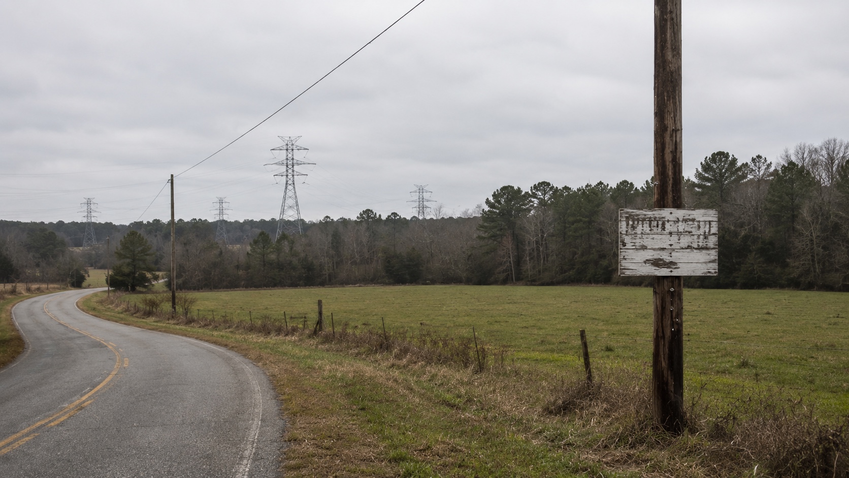 A two-lane country road in rural Alabama, an unassuming roadside pasture in the middle distance, and the faint silhouette of high-voltage transmission towers crossing the horizon — a hint that this land is being assembled because of its proximity to power infrastructure.