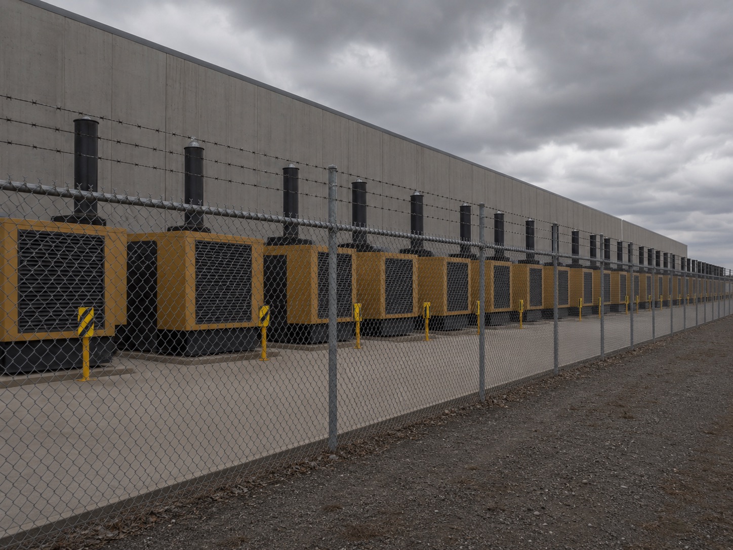 A long row of industrial diesel backup generators behind a chain-link security fence with three-strand barbed wire, against a massive concrete data center wall.