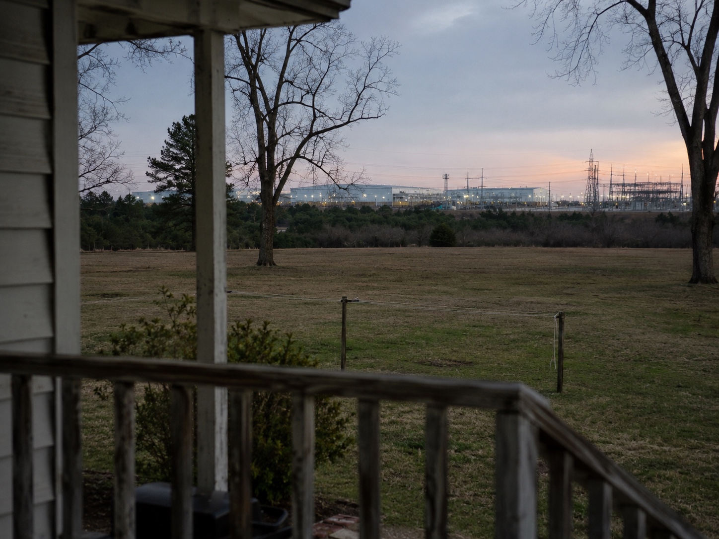 The view from the porch of a modest rural Alabama home; a hyperscale data center campus, transmission towers, and bright industrial security lighting visible on the horizon at dusk.