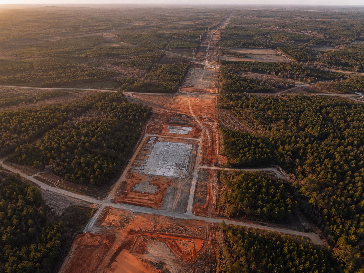 Aerial of a continuous Black Belt forest cut by industrial access roads, a transmission-line right-of-way, and the cleared concrete pad of a single new building.